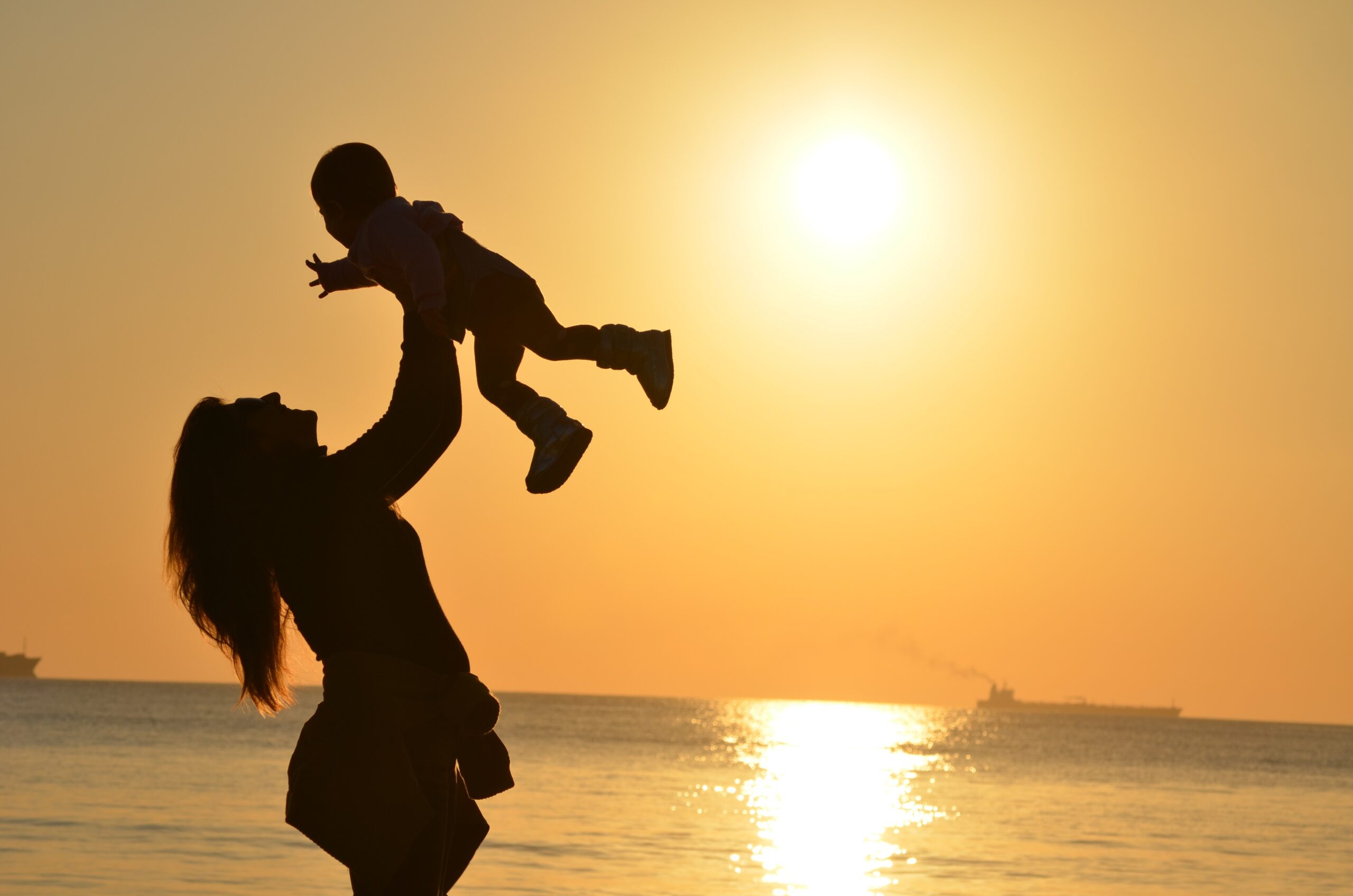 Woman and child standing in front of water while sun is setting